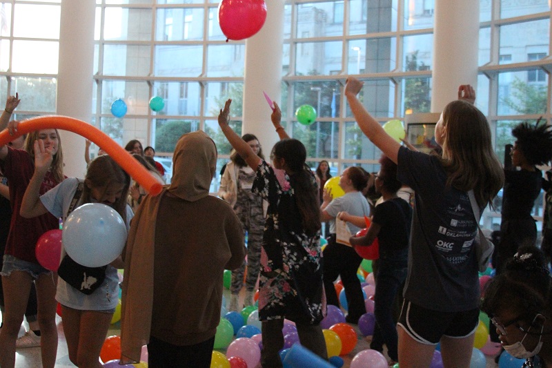 A group of teens dance and wave their hands in the air as balloons drop from the ceiling.